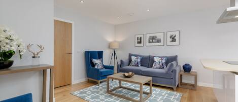 A modern living room with a grey sofa, blue armchair, and a coffee table. There is a wooden door, a small side table with a vase, and a blue rug.