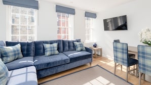 A modern living room with a blue sectional sofa, a flat-screen TV mounted on the wall, and a dining area with a small table and chairs. - Destiny Scotland Royal Mile Residence (Edinburgh)