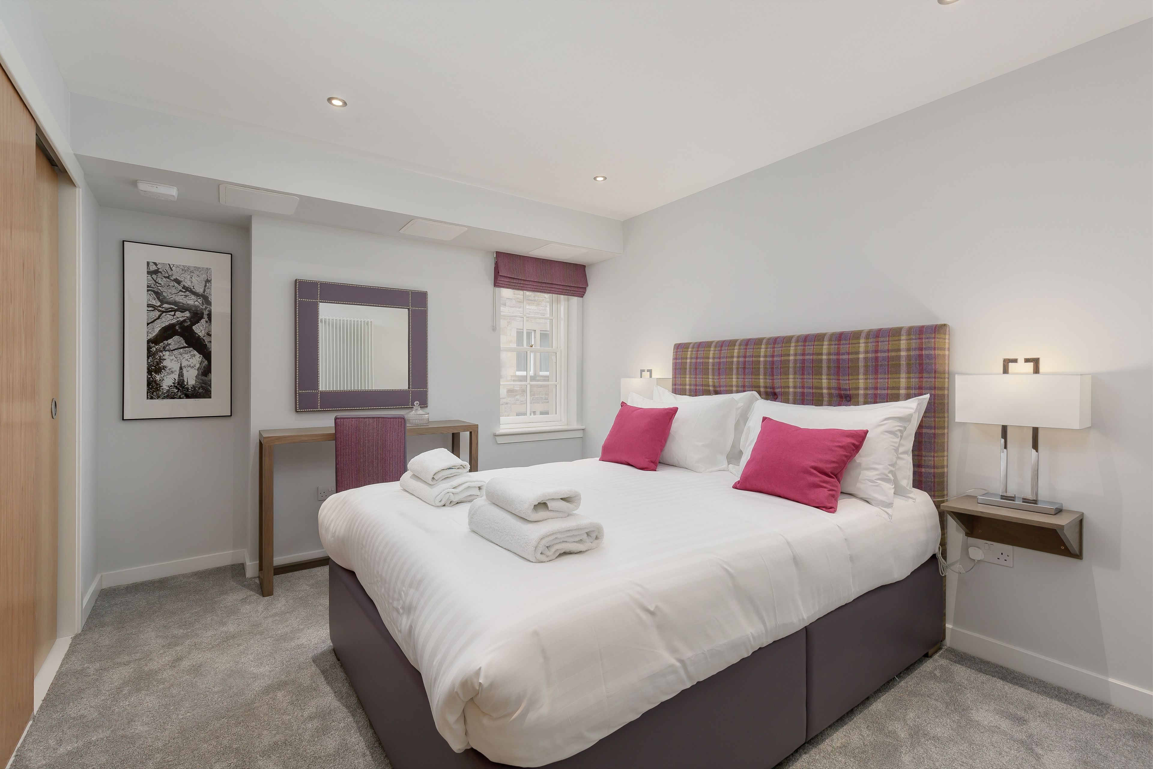 A modern bedroom with a large bed, a bedside table with a lamp, a window with a view, and a framed picture on the wall.