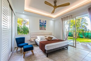 Quarto com cama, cadeira, ventoinha no teto e janela com vista para o verde.