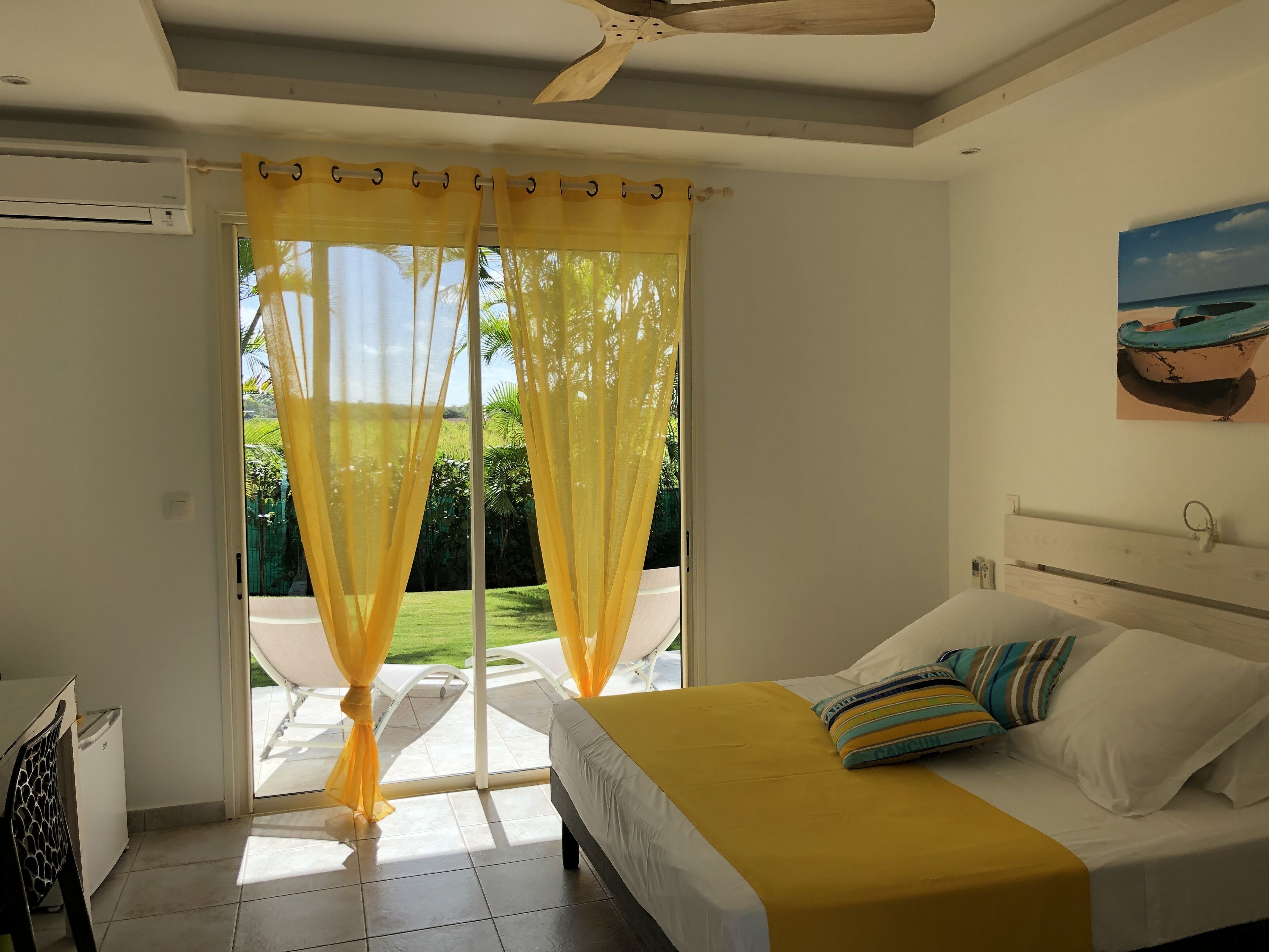 A bedroom with a bed, yellow curtains, a ceiling fan, and a view of a garden.