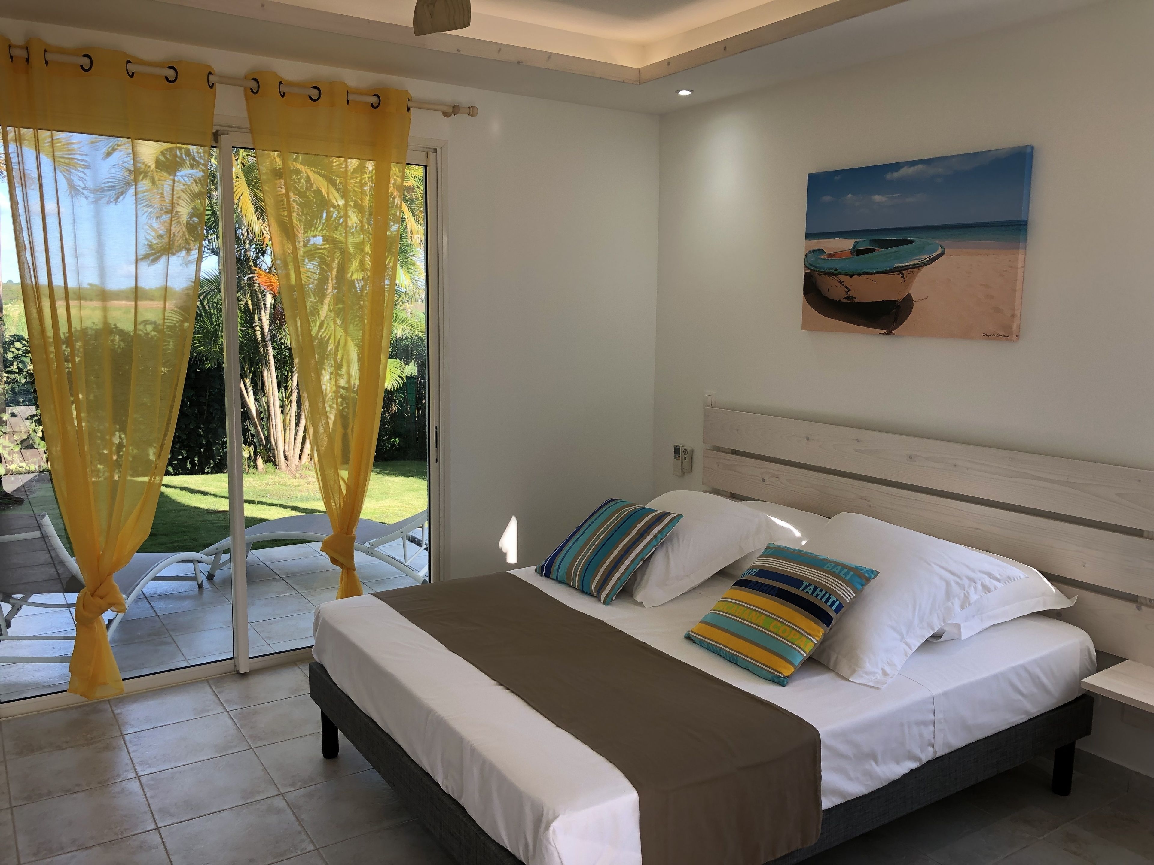 A bedroom with a bed, a painting of a beach scene, and a view of a garden through a glass door.