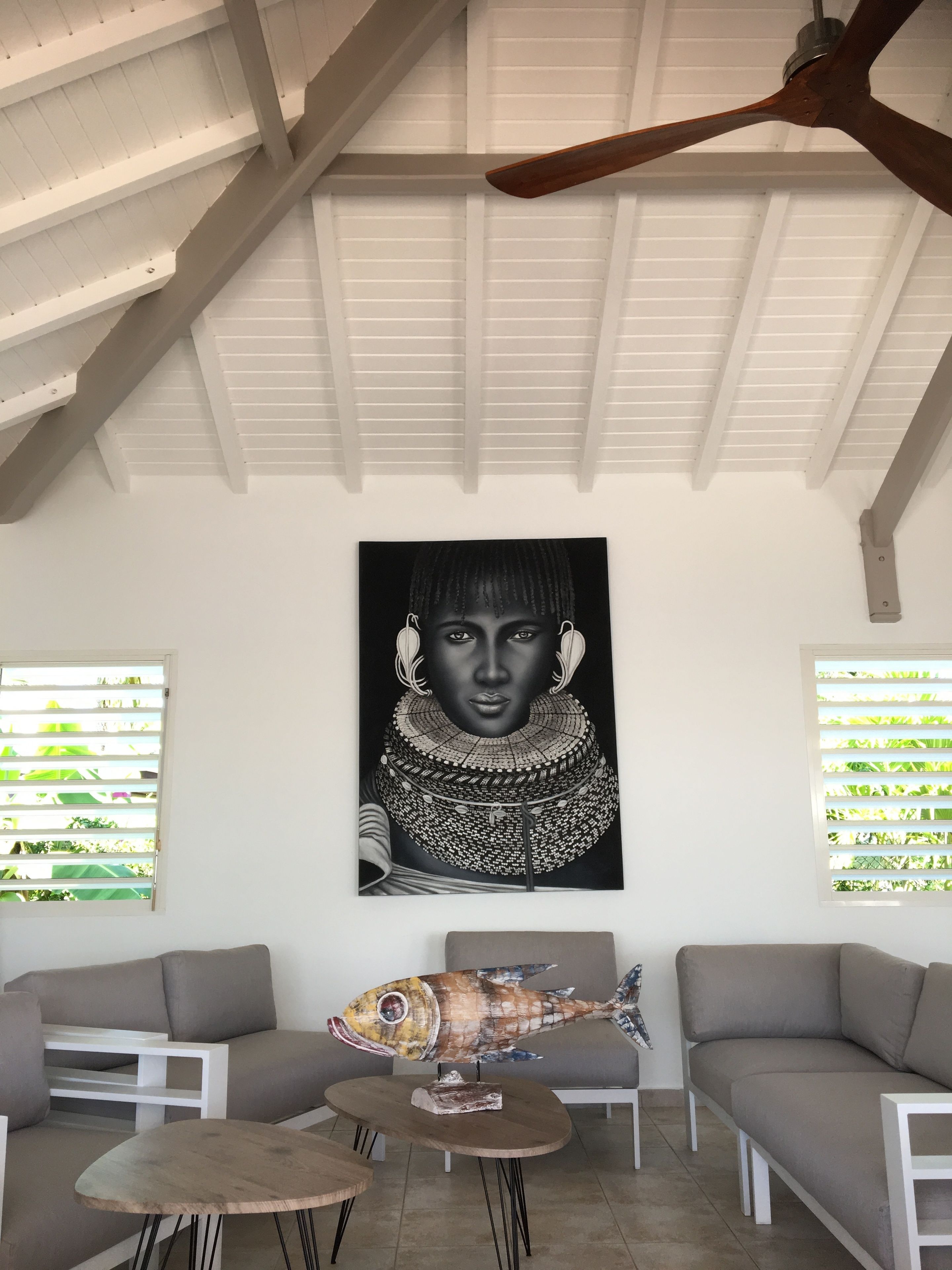 Una habitación con techo alto, paredes blancas y un ventilador de techo. Se observa una gran obra de arte blanco y negro en la pared, una escultura de pez sobre una mesa y dos sofás grises.