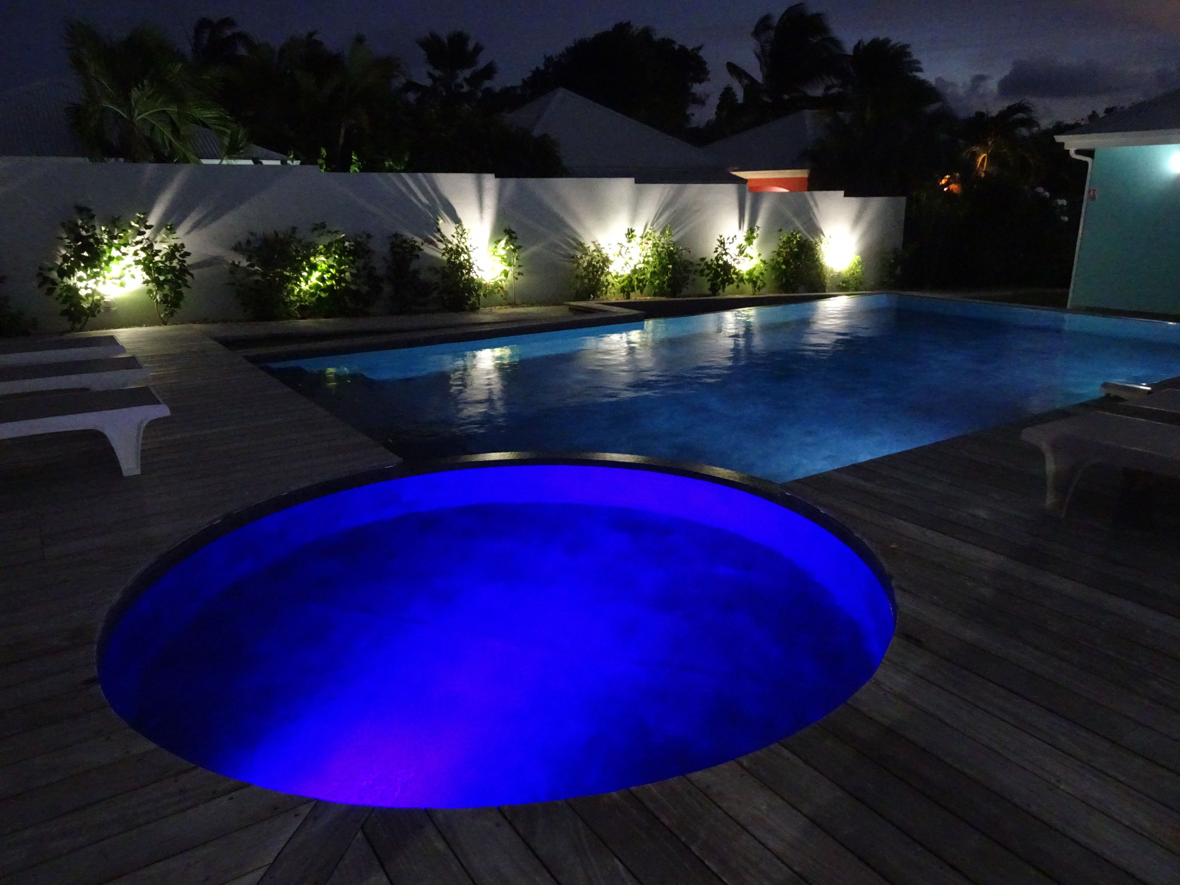 A swimming pool with a blue-lit hot tub, surrounded by a wooden deck and illuminated plants.