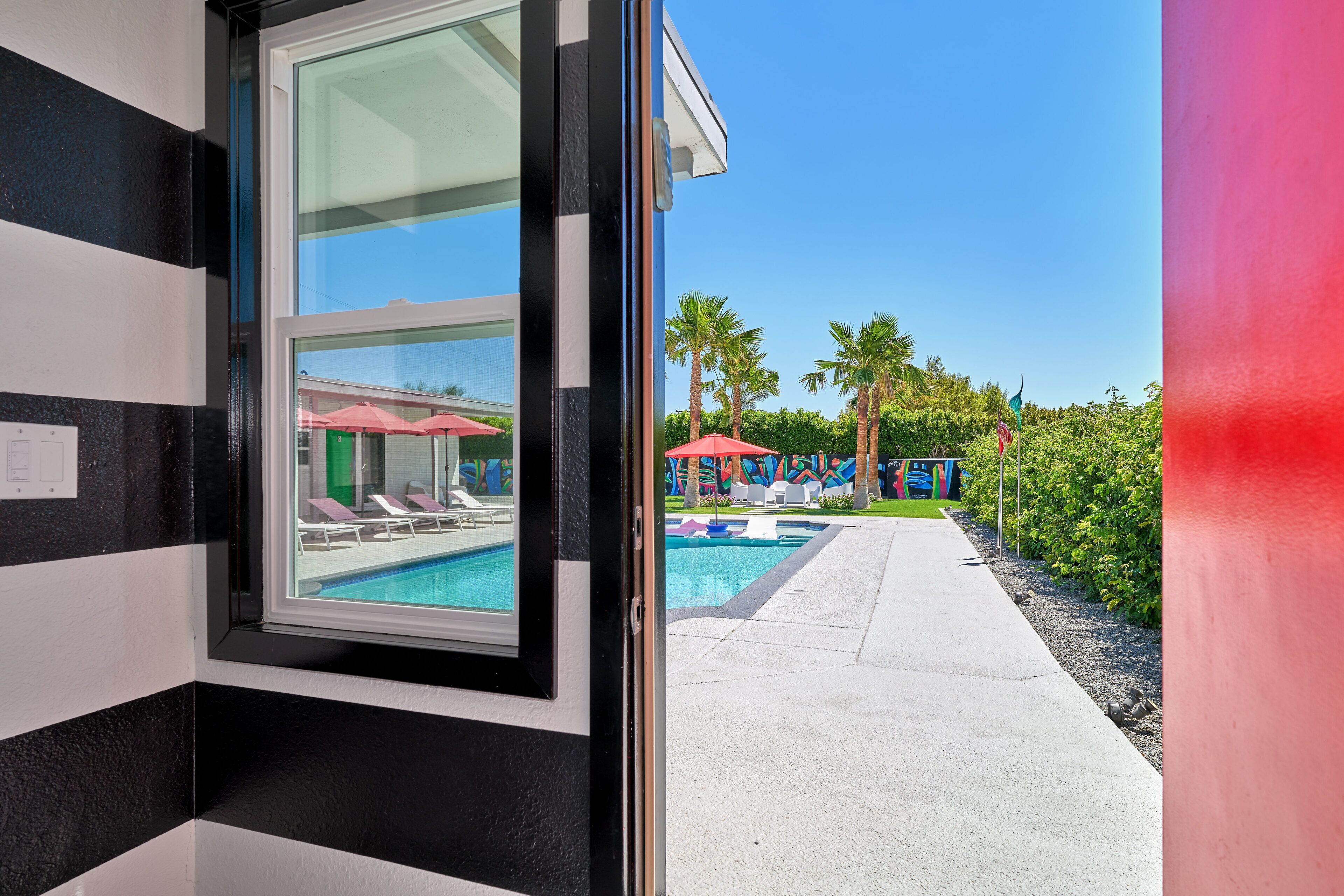 A modern hotel room with a large window, a pool area with lounge chairs and umbrellas, and a clear blue sky.