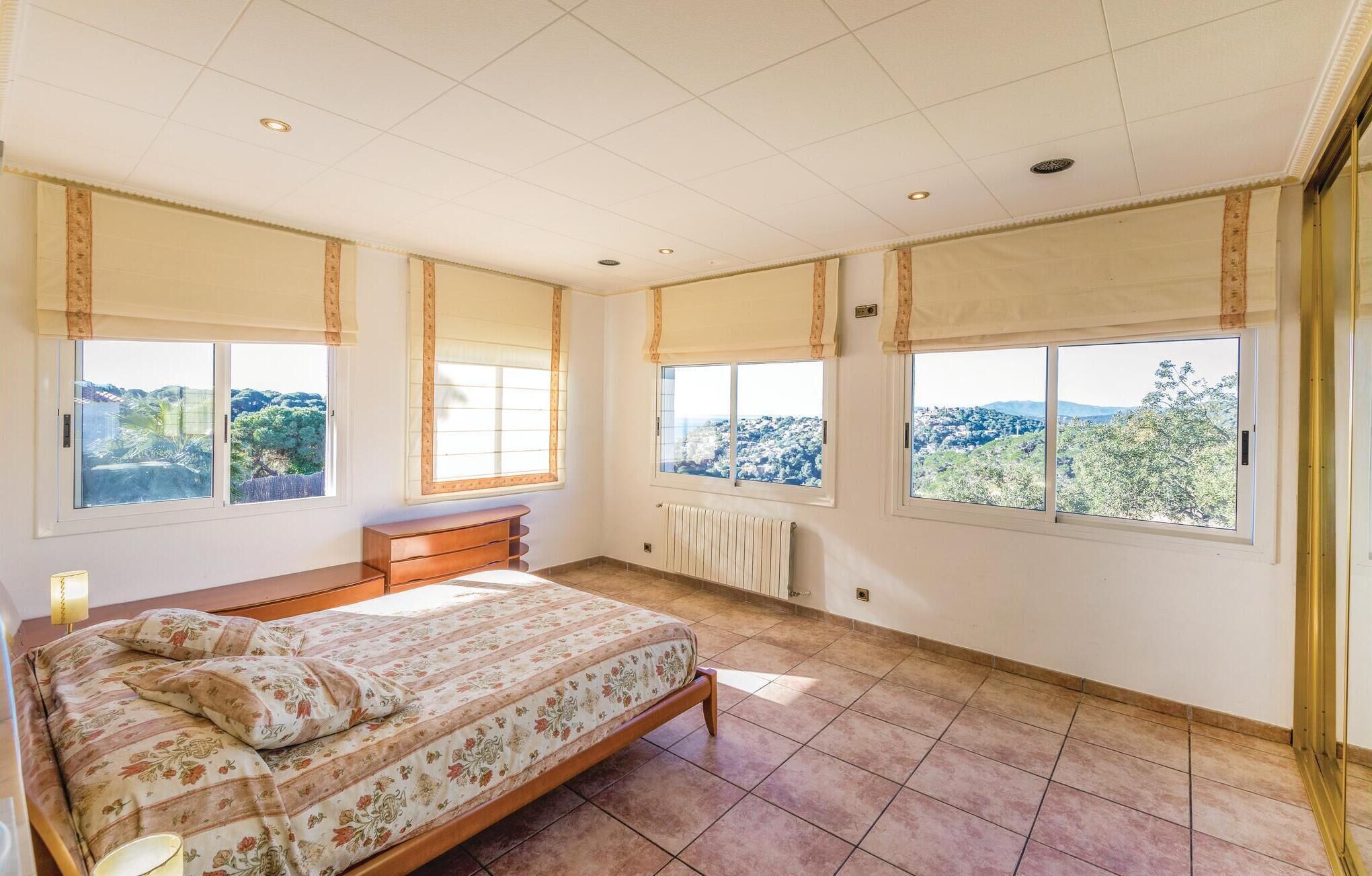 Ein Schlafzimmer mit einem großen Bett, zwei Fenstern mit Vorhängen, einem Heizkörper und Blick auf eine hügelige Landschaft.