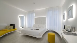 A modern hotel room with a curved bed, a yellow ottoman, a white desk with a coffee maker, and a large window with sheer curtains. - AHD Rooms (Milan)