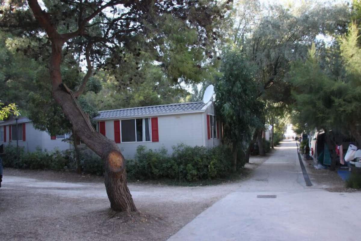 A row of small, single-story buildings with red shutters, surrounded by trees and a gravel path.