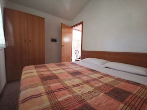 A bedroom with a bed, a wooden wardrobe, and a door leading to another room.
