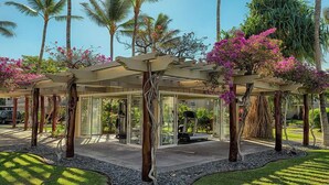 A covered outdoor area with a glass enclosure, surrounded by palm trees and flowering plants.