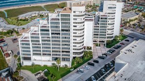 A high-rise hotel with multiple balconies, a parking lot, and a view of the beach and ocean.