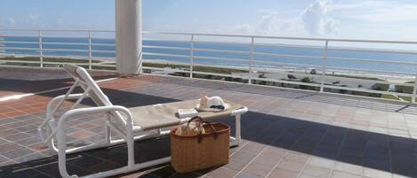 A sun loung Chair and table on a tiled deck with a view of the ocean and a road.