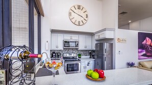 A modern kitchen with white cabinets, a stainless steel refrigerator, and a microwave. There is a clock on the wall and a wine bottle on a cart.