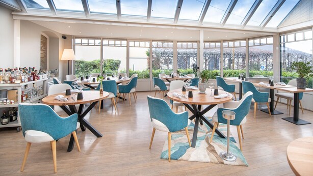 Un área de comedor moderna con mesas redondas y sillas color turquesa, grandes ventanales con vistas a la vegetación y una zona de bar con estantes llenos de botellas.