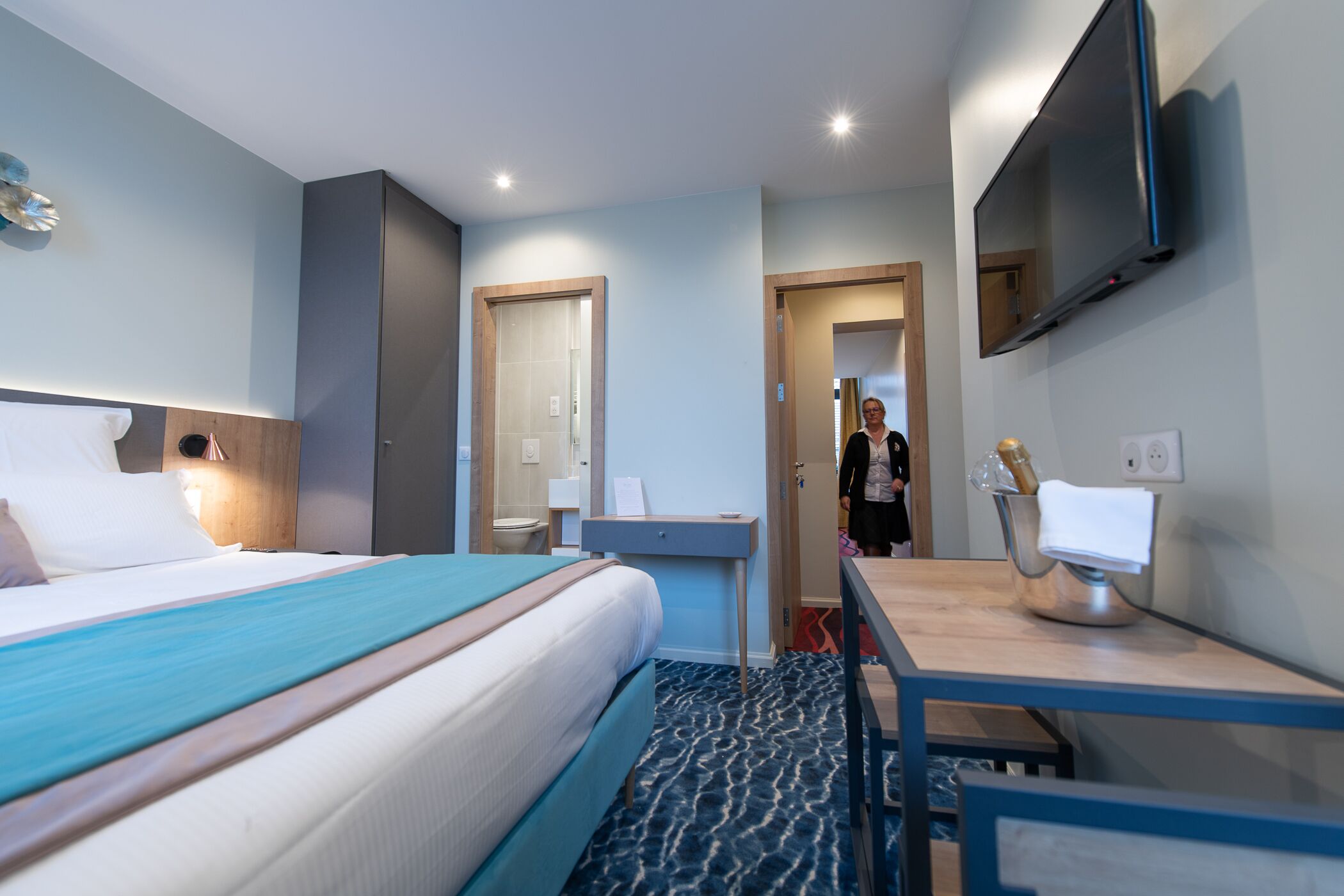 Habitación de hotel con cama, mesita de noche, televisión y baño visible a través de una puerta abierta.