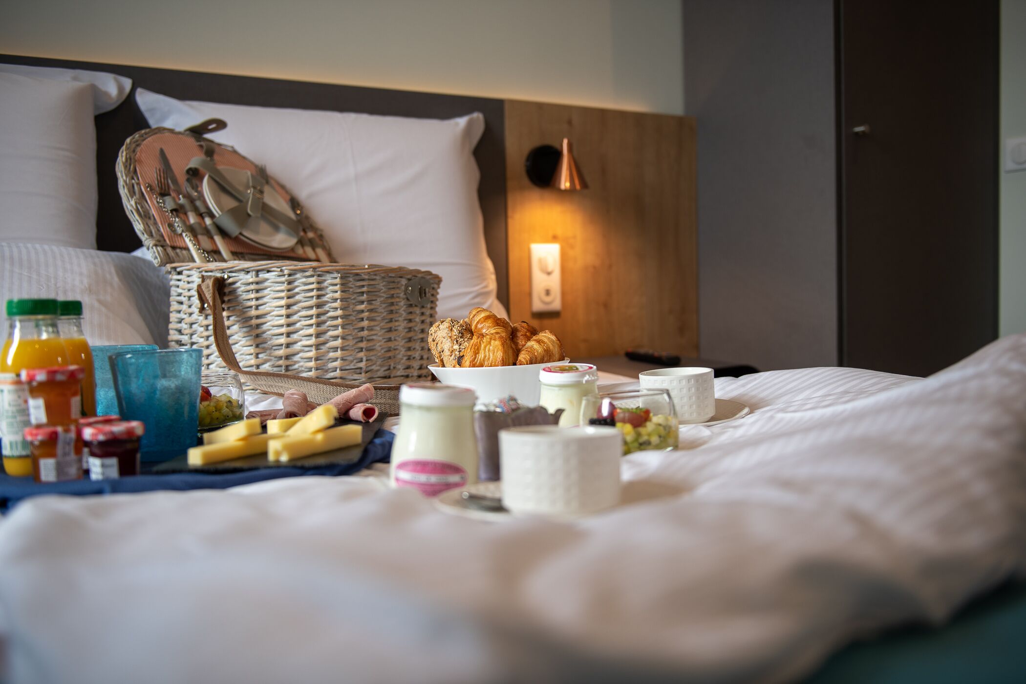 Een keurig opgemaakte ontbijtbuffet op een bed, met een rieten mand, croissants en diverse ontbijtartikelen.
