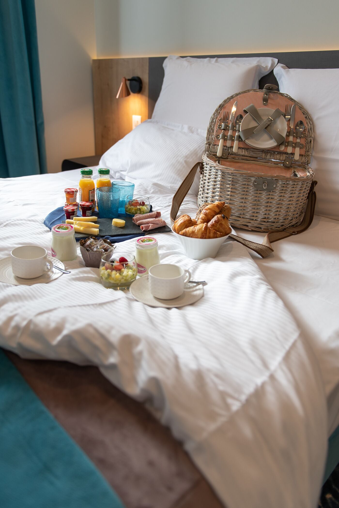 A neatly made bed with breakfast items including croissants, fruit, and beverages, and a wicker basket on the side.