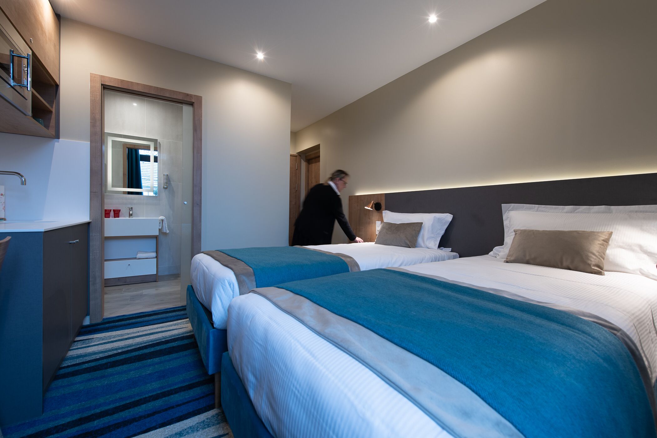 A modern hotel room with two beds, a bathroom, and a small desk area.