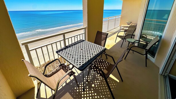 A balcony with a table and chairs, overlooking a beach and the ocean.
