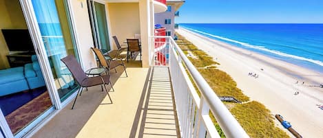 A balcony with a view of the beach, outdoor seating, and a pool area.