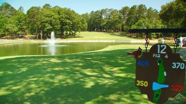 Un parcours de golf avec un étang, une horloge affichant les scores et une personne visible au loin.