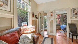 A cozy living room with a leather sofa, a wooden coffee table, and a large window with a view of trees. There are framed pictures on the walls and a door leading to another room. - Near the Ocean/Self Check-in/Out/Wifi/High Cleaning Standards  (Victoria)