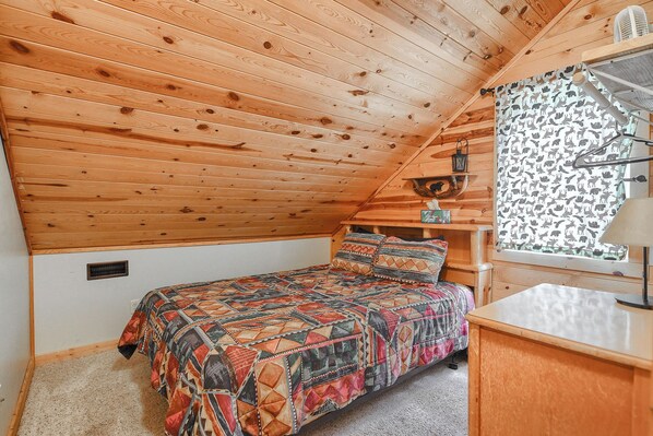 A small, cozy room with a sloped ceiling, a bed with a patterned quilt, a wooden desk, and a window with patterned curtains.