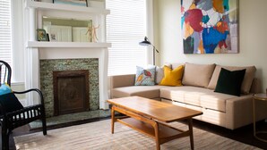 A living room with a beige sofa, a wooden coffee table, a fireplace, and a colorful abstract painting on the wall. - The Calliope, Historic Corner Apartment in the Heart of Downtown. (Savannah)