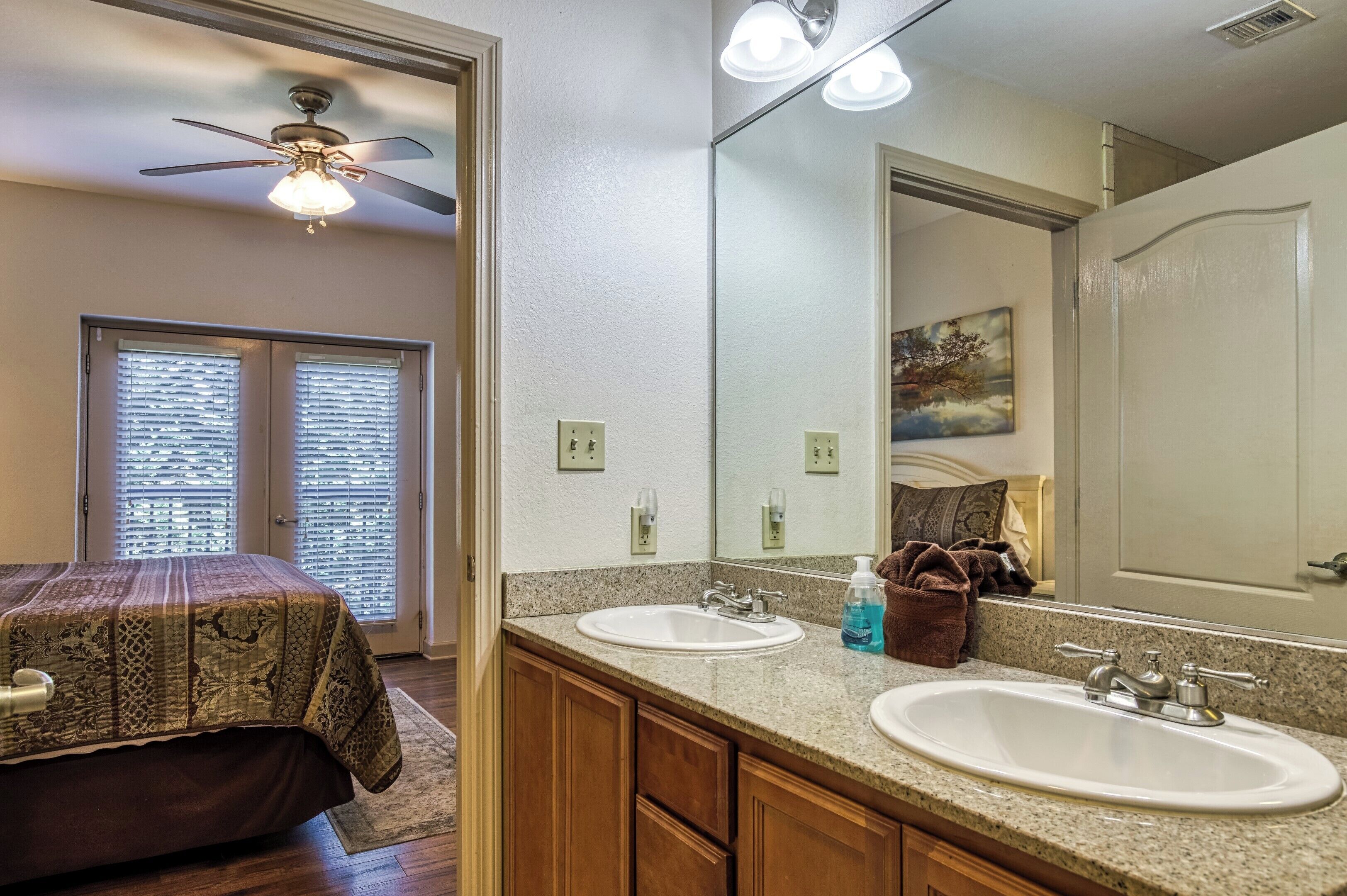 A bedroom with a bed, two windows with blinds, a ceiling fan, a bathroom with two sinks, a mirror, and a painting on the wall.