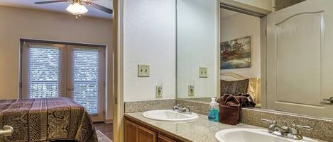 A bedroom with a bed, two windows with blinds, a ceiling fan, a bathroom with two sinks, a mirror, and a painting on the wall.