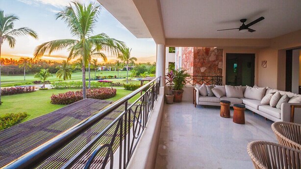 A balcony with a sofa, chairs, and a railing overlooking a landscaped garden with palm trees and a sunset view.
