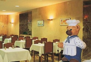 Un espace repas avec des tables et des chaises, une grande peluche en forme d’ours et des luminaires fixés au mur.