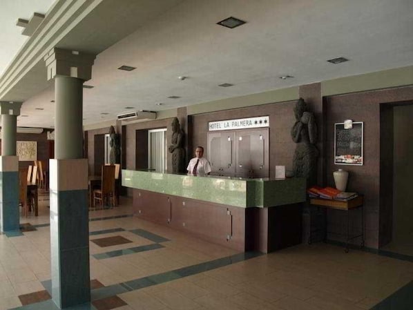 Un espace d’accueil d’hôtel comprenant un comptoir de réception, un réceptionniste et une enseigne au-dessus du comptoir indiquant « HÔTEL LA PALMERA ».