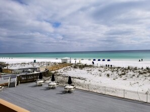 A beach view with a wooden deck, outdoor seating, and a sandy beach area.