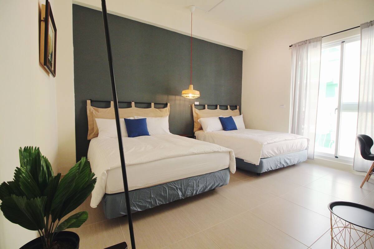 Habitación de hotel con dos camas, una planta en maceta, una mesa de comedor y un amplio ventanal con cortinas.
