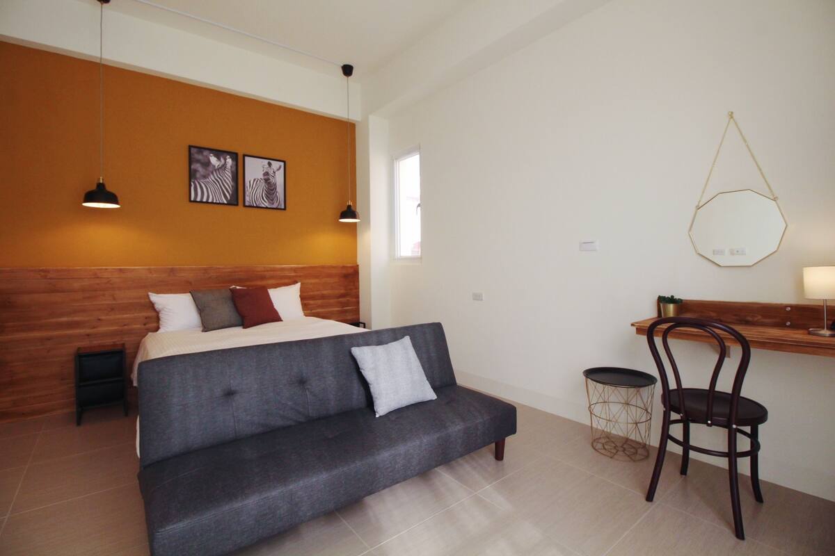 Un dormitorio con cama, un sofá gris, cabecera de madera, escritorio con silla y lámpara colgante.