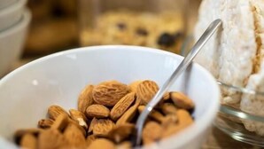 A white bowl filled with almonds on a wooden surface, with a glass jar and a spoon in the background.