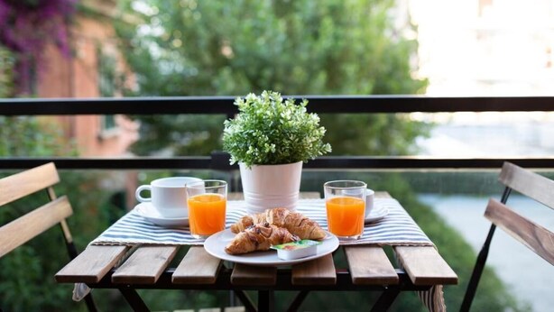 A wooden table with a plate of croissants, a cup, and two glasses of orange juice, a small potted plant, and a view of greenery and buildings.