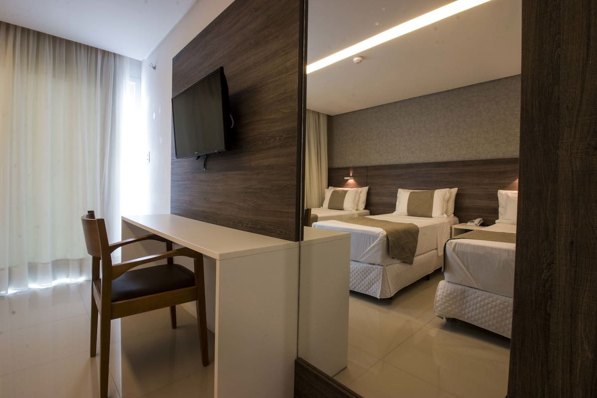 A hotel room with a wooden desk, two single beds, a TV mounted on the wall, and a large mirror.