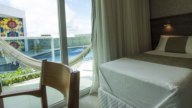 Habitación de hotel con cama, una silla, una hamaca y vistas a una piscina y un edificio con diseños coloridos.