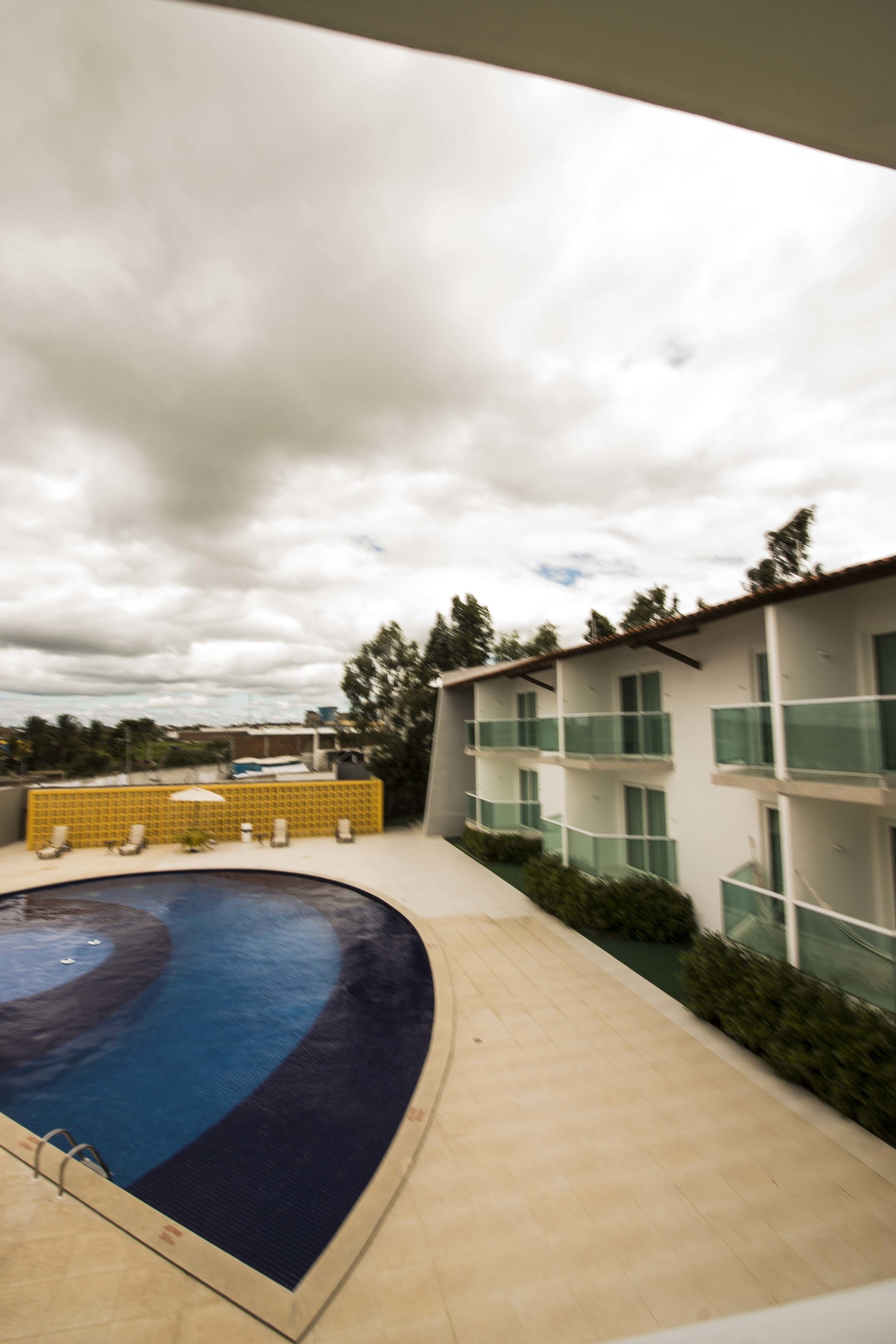 A swimming pool with a curved edge, surrounded by a tiled deck. There are yellow chairs and a white umbrella near the pool.