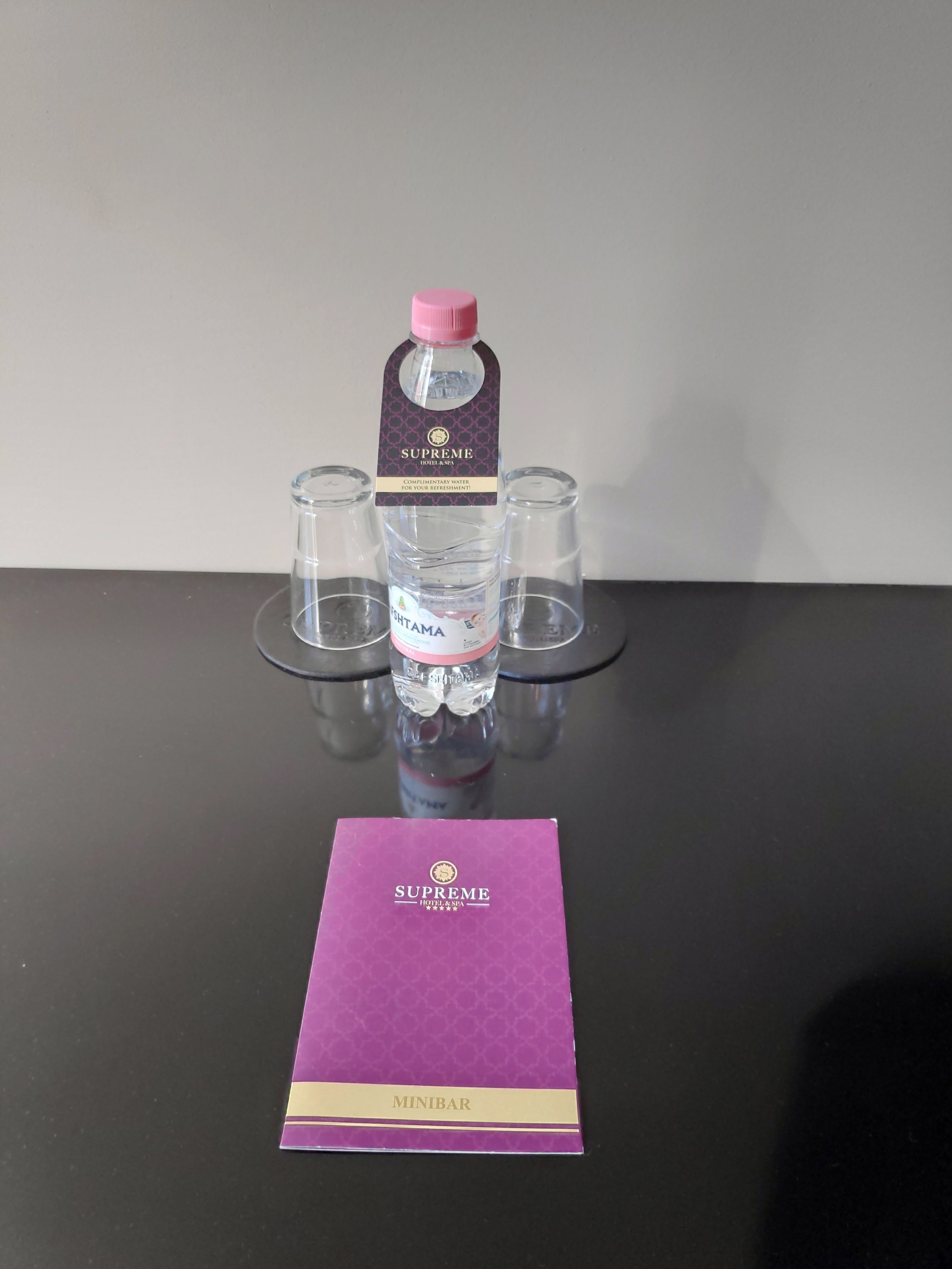 Uma garrafa de água com o rótulo "Supreme Hotel & Spa", uma etiqueta, três copos vazios e um livreto roxo intitulado "Minibar".