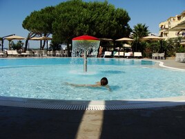 Una piscina con fuente, sillas de descanso y sombrillas.