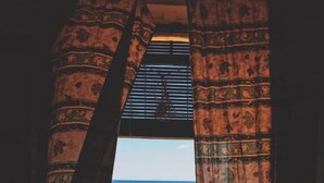 Une vue à travers une porte, encadrée de rideaux à motifs, qui donne sur un balcon avec une balustrade d’où l’on aperçoit la mer.