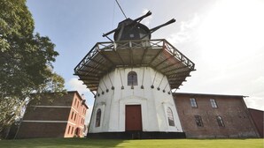 Historiallinen tuulimylly, jossa on valkoinen julkisivu ja punainen ovi, ja sen ympärillä on tiilirakennuksia ja nurmialue.