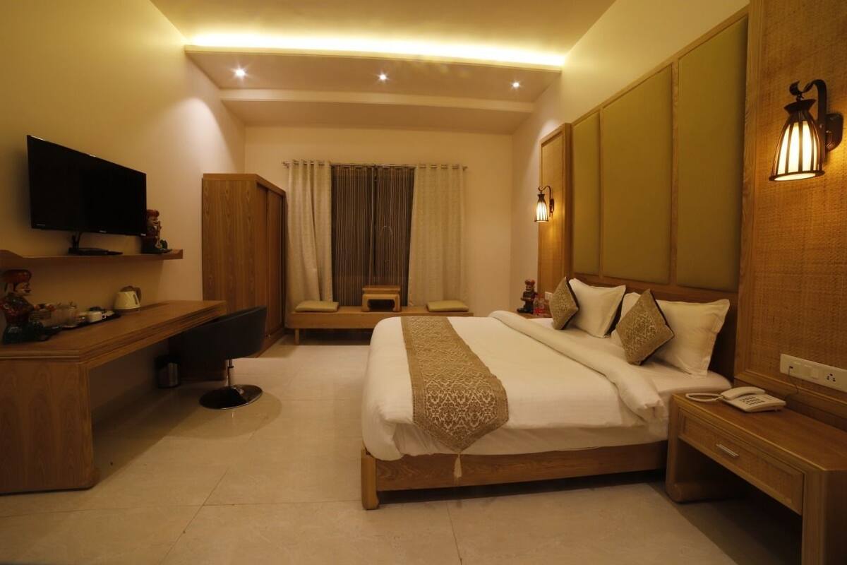 Quarto de hotel com cama, mesa de cabeceira, luminária, televisão, escrivaninha e cortinas.