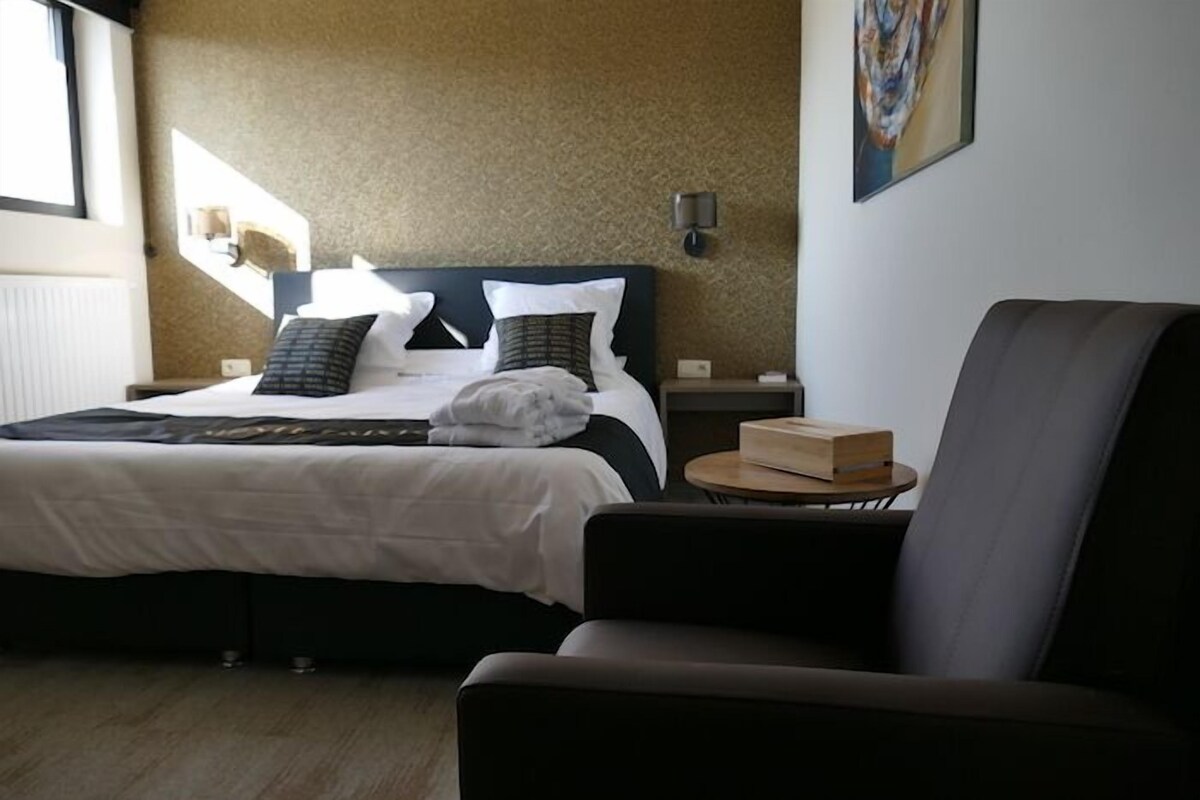 A hotel room with a bed, a chair, a bedside table, and a painting on the wall.