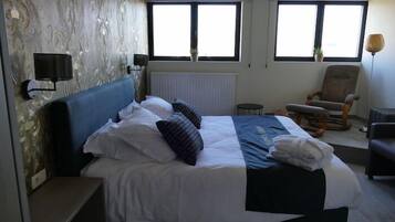 A hotel room with a large bed, two windows, a chair, and a small table.