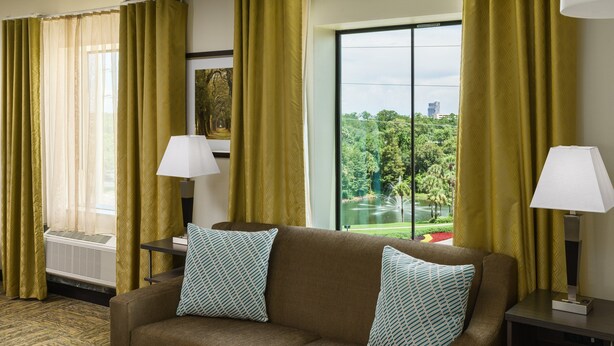 A hotel room with a brown sofa, two patterned pillows, a side table with a lamp, and a large window offering a view of a park and buildings. - Candlewood Suites Orlando - Lake Buena Vista by IHG (Orlando)