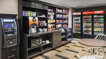 Zona de autoservicio con cajero automático, máquina de café, estantes de snacks y dispensadores de bebidas.